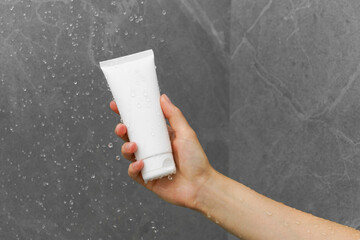 Female hand holding a white mockup of a tube of body wash or face moisturizer under water drops in...