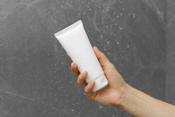 A woman's hand holds a white Mockup tube of face cream or gel under water drops in the shower. The...