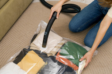 A woman uses a vacuum cleaner to extract air from a transparent vacuum bag with clothes while sitting on the floor. The concept of compact storage and saving space in the closet