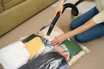 A young woman uses a vacuum cleaner to extract air from a vacuum bag with clothes for compact storage in a closet while sitting on the floor. Space saving concept, compressed packaging and seal bag - obrazy, fototapety, plakaty