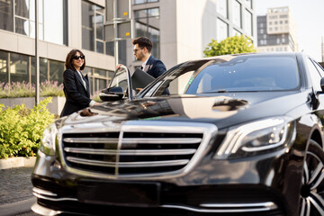 Female chauffeur helps a businessman with laptop to get out of the car, opening a door. Concept of personal driver, luxury taxi or business trips