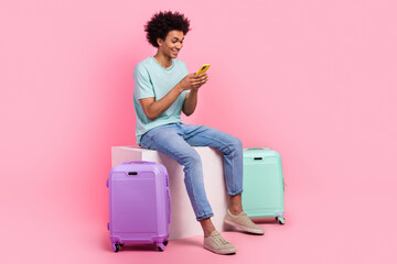 Full size photo of nice young guy device sit white cube travel baggage wear trendy blue outfit isolated on pink color background