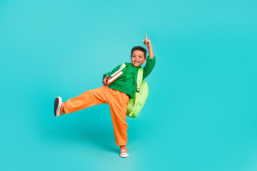 Full size photo of small boy with rucksack wear green hoodie hold book directing up at empty space isolated on turquoise color background