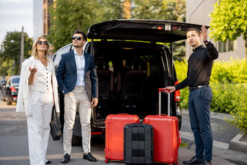 Business couple standing by a minivan taxi waiting for their chauffeur or porter to help them with a suitcases. Concept of business trips and travel - 700331291