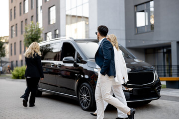 Business people walk to minivan taxi from office building, female driver waits for them nea...