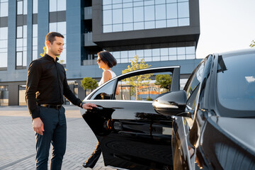 Chauffeur helps an elegant business woman gets in car, opening door of a premium taxi near office building or hotel. Concept of personal driver or business trips