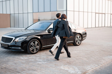 Businessman and businesswoman walk together to car on parking lot near modern building, going to drive by luxury car