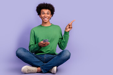 Full length photo of cheerful cute man dressed green sweater texting modern device pointing empty space isolated violet color background