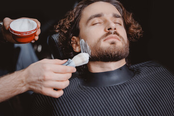 Barber applies shaving foam with brush to man beard in barbershop, warm toning, top view.