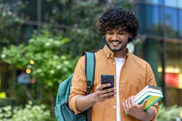 Indian smiling young male student standing outside with backpack and books and using mobile phone
