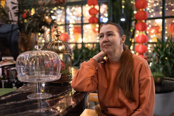 Middle-aged woman wearing peach fuzz coloured clothes sits in cafe decorated for Christmas and Chinese New Year. Chinese lanterns, glowing garlands. Festive mood. Self-love, self-care. Mental health