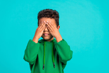 Portrait of funny good mood small boy with curly hairstyle wear green hoodie palms cover eyes...