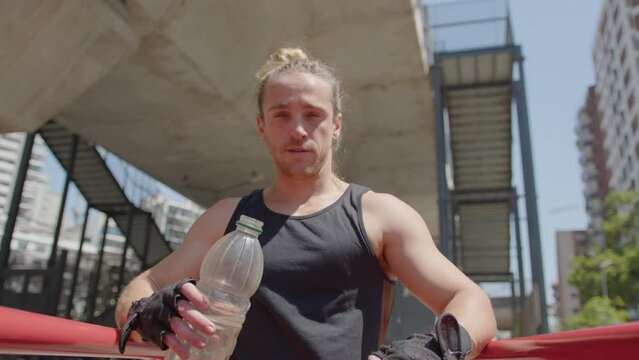 Muscular man in sportswear holding bottle of water, smiling and posing for camera at parallel bars after outdoor workout. Zoom shot, video portrait