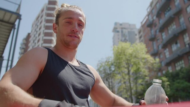 Strong sportsman holding bottle of water and posing for camera by parallel bars at outdoor gym after training. Zoom shot, video portrait