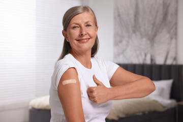 Senior woman with adhesive bandage on her arm after vaccination showing thumb up indoors