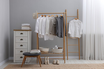 Wardrobe organization. Rack with different stylish clothes, ottoman and chest of drawers near grey wall in room