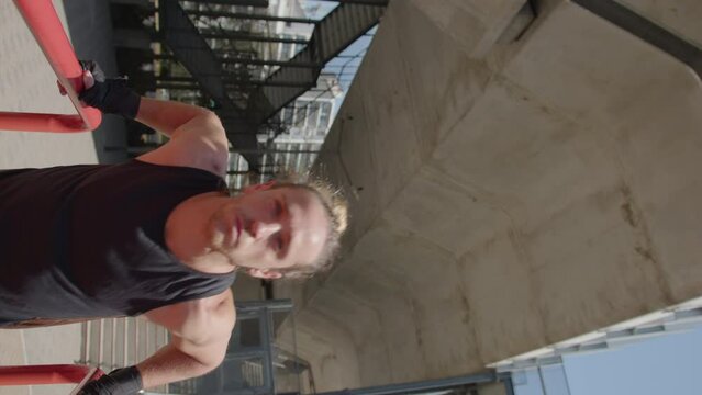 Athletic man in sportswear and gloves doing tricep dips exercise on parallel bars during street workout outside. Vertical format video