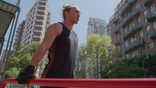 Strong male athlete in sportswear practicing tricep dips on parallel bars at outdoor gym while having street workout in the city. Handheld camera shot