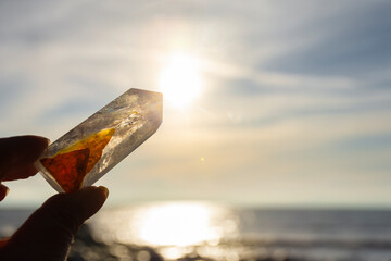 A close up image of a hand holding a clear quartz crystal tower against a colorful sunset with an...