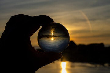A silhouette image of a hand holding a glass lens ball at sunset with an glowing orange sky.