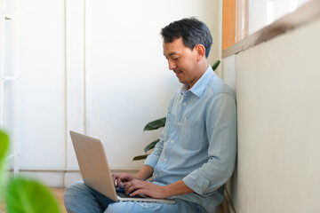 Fototapeta premium The young man asian is working from home in the living room, using his mobile phone and computer to command his work remotely.