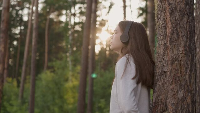 Woman with headphones leans on tree trunk in forest. Young lady listens to melancholy music thinking about bitter experience at sunset. Feeling of solitude