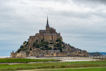 View of Mont Saint Michel Abbey Castle in Normandy France under a cloudy sky with grassy banks in the foreground