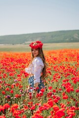 Naklejka na ściany i meble Happy woman in a poppy field in a white shirt and denim skirt with a wreath of poppies on her head posing and enjoying the poppy field.