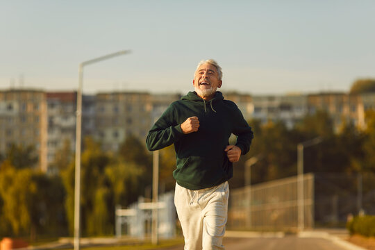Cheerful fit senior man enjoying jogging workout. Portrait happy smiling handsome bearded 60 - 70 year old man running on city street on sunny summer morning. Sport, fitness, healthy lifestyle concept
