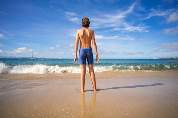 A teenager is standing on the sandy shore of the ocean.