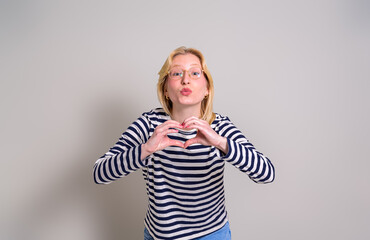Portrait of loving young woman blowing kiss and forming heart shape over isolated white background