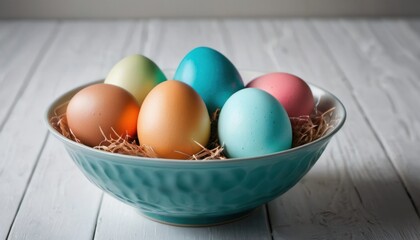  a blue bowl filled with colored eggs on top of a white wooden table next to a white painted wall and a white painted wall behind the bowl is filled with straw.