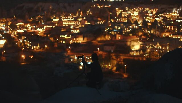 Young man filming stars with a camera and a tripod at night. City lights in the background.