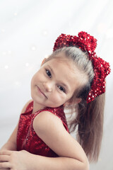 Obraz na płótnie Canvas christmas portrait of a smiling happy little girl in red dress with glitter ribbon on head, light background