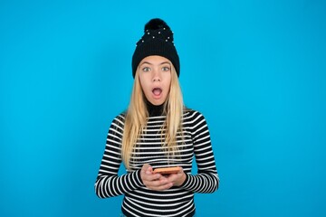Shocked beautiful caucasian teen girl wearing striped sweater opens mouth hold phone reading advert...