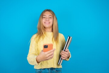beautiful caucasian teen girl wearing yellow sweater holds mobile phone in hands and rejoices positive news, uses modern cellular