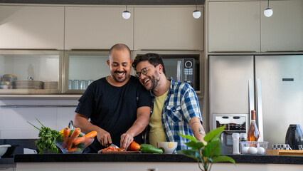 Happy male gay couple standing in kitchen at home preparing together yummy dinner on first dating,...