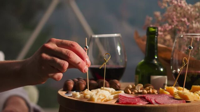 Man holds cheese on skewer at low coffee table closeup. Assortment of delicious appetizers on small wooden table for gourmets. Snack plate to enjoy wine