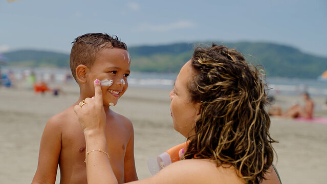 mother applying sunblock lotion on sons skin for sun protection little boy getting ready to swim on the beach with mom using sunscreen caring for childs health on sunny day 
