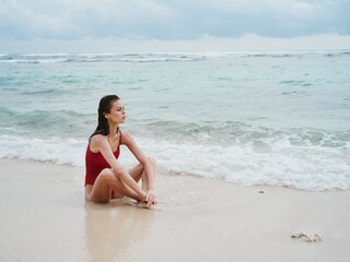 Fototapeta na wymiar A young woman sitting by the sea in a red bathing suit on the sand near the water and looking at the horizon 