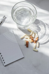 Blank paper sheet card, pills and capsules, glass with water on white marble table. Medical prescription or health care planning