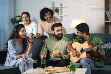 group of happy friends enjoying by playing guitar and singing during home gathering - concept of...