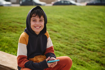 Confident portrait of a multi ethnic preteen boy in stylish hoodie, holding mobile phone, smiling...