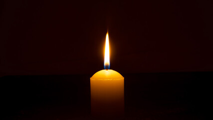 A single burning candle flame or light glowing on a big yellow candle isolated on red or dark background on table in church for Christmas, funeral or memorial service