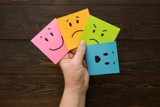 Hand holds notes with a basic emotions - joy, sadness, anger, fear or surprise. Four colorful squares with emotions on a brown wooden background