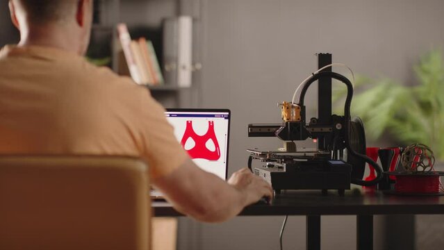 A man graphic designer develops a model of postoperative fixation bandage in red colour for the heart and chest for women, depicted on a laptop screen. A 3d printer machine works in automated