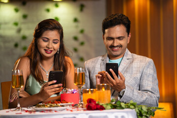 Indian couples at romantic candlelight dinner busy using mobile phone at restaurant - concept of...