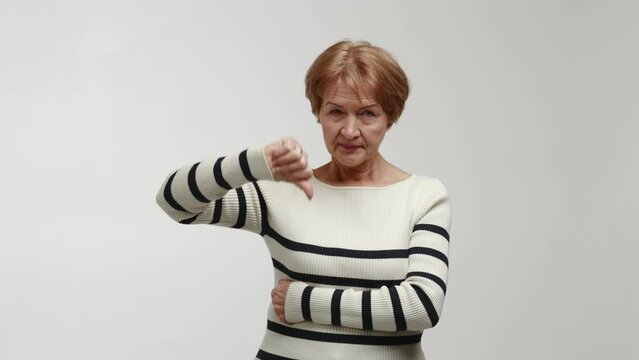 An elderly strict and serious woman stands and expresses emotions of discontent, disapproval and inadmissibility. Gesture of fist with thumbs down, demonstration of categorical rejection, condemnation