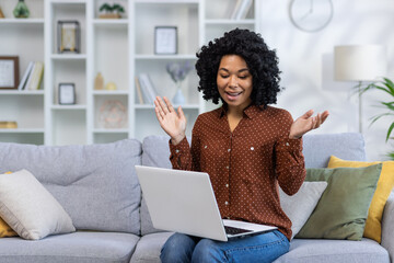 African American young woman sitting on sofa at home and talking smiling and emotional on video...