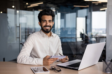 Portrait of a young Indian successful man smiling and looking at the camera, sitting in the office...